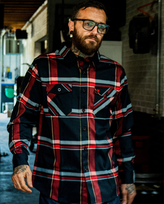 Man wearing a plaid red and black flannel shirt with tattoos on arms, standing outdoors - The Devil in the Details Flannel from Greasy Hands Society