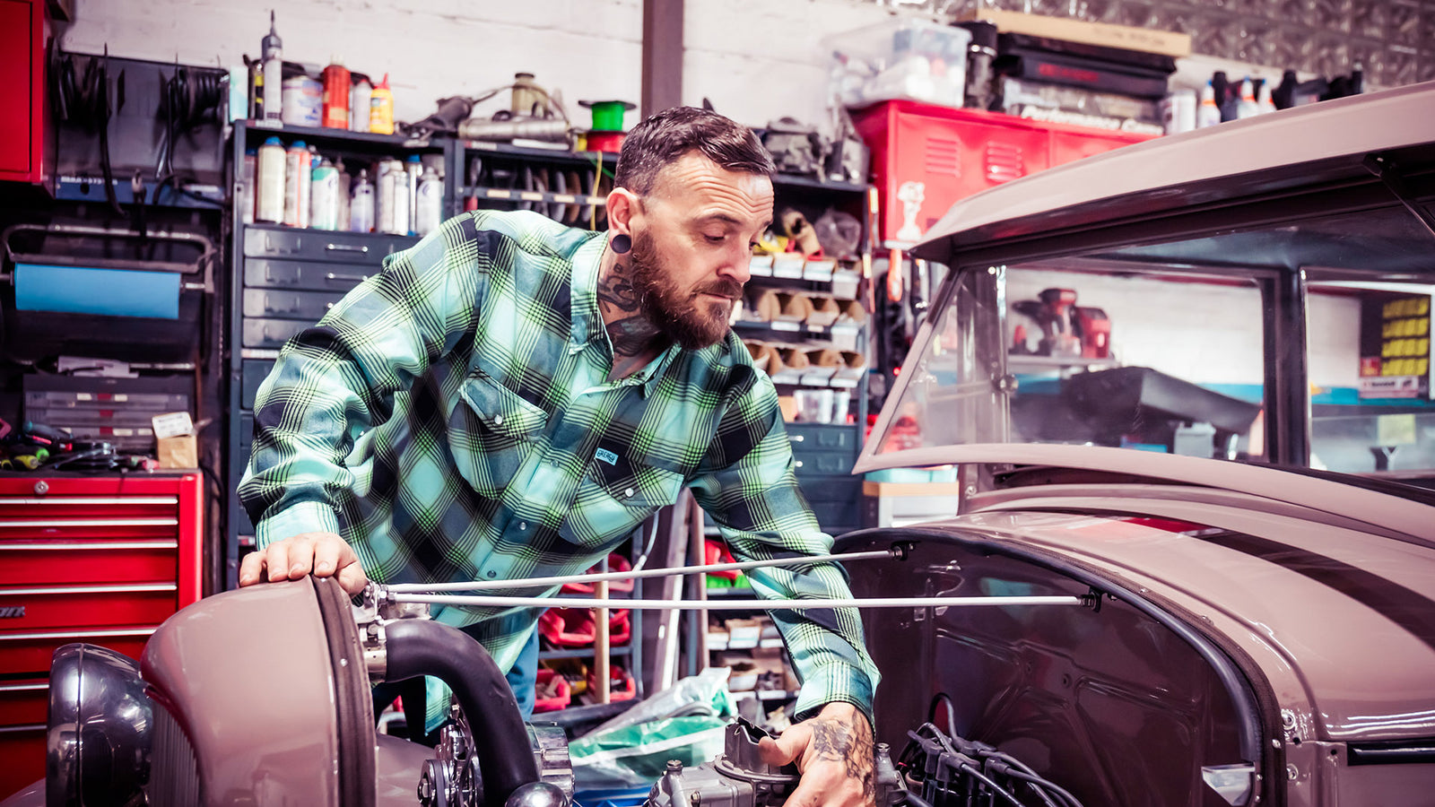man working on a car in the greasy hands society sucker punch blue and green and black flannel.
