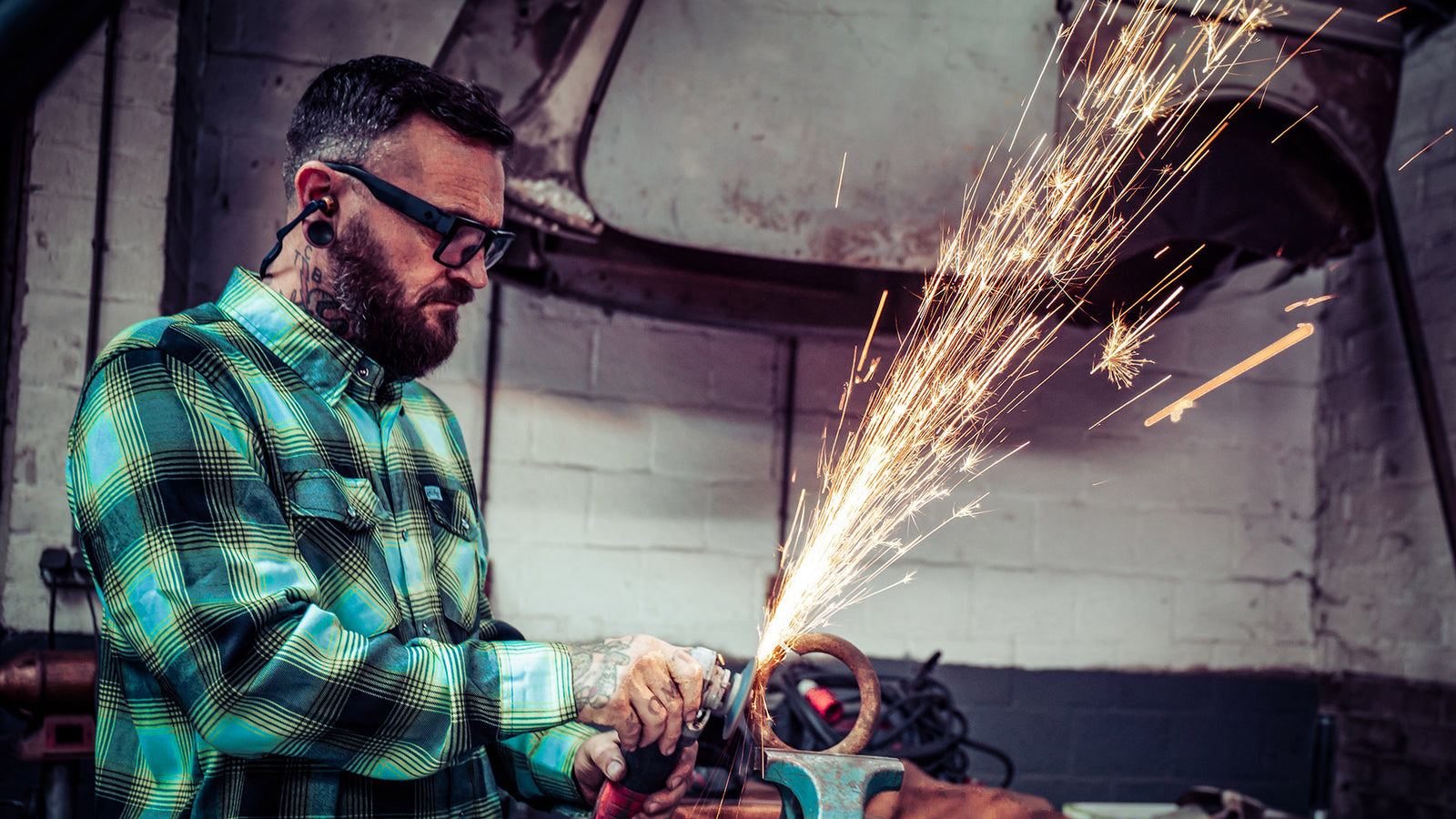 Man using a grinder with sparks flying in an industrial setting in the greasy hands society sucker punch blue and green and black flannel.