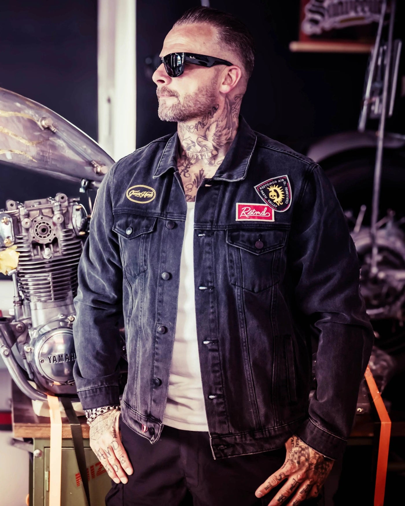 Man wearing a denim jacket with patches in a garage setting - The Black Denim Jacket by Greasy Hands Society