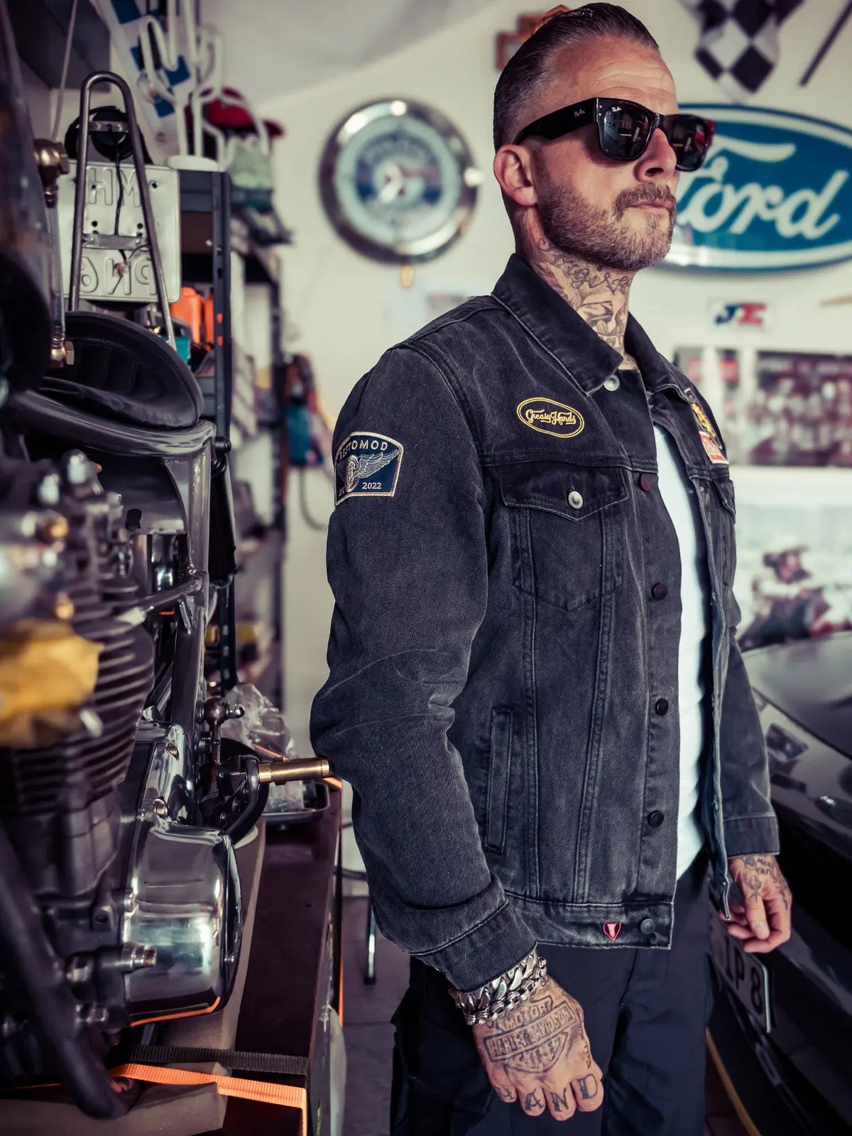 man with tattoos and sunglasses wearing a black denim jacket in a industrial environment and motorcycle shop - The Black Denim Jacket by Greasy Hands Society