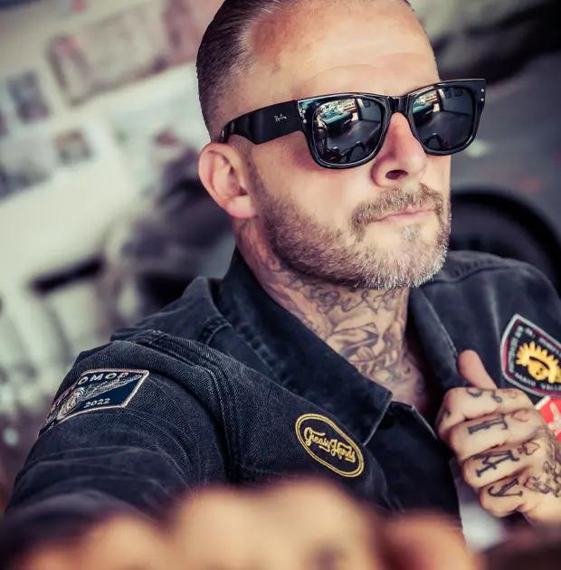 A close-up lifestyle shot of a man wearing black sunglasses and a black denim jacket with embroidered patches, including one that reads 'Restomod'- The Black Denim Jacket by Greasy Hands Society
