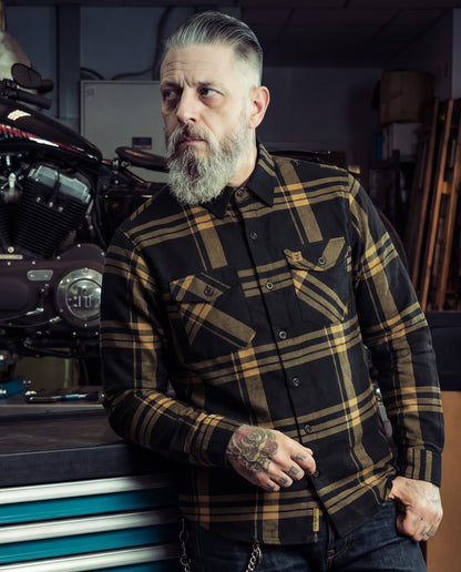 Man wearing a plaid black and gold flannel shirt in a workshop setting with motorcycles - The Two-Lane Flannel from Greasy Hands Society