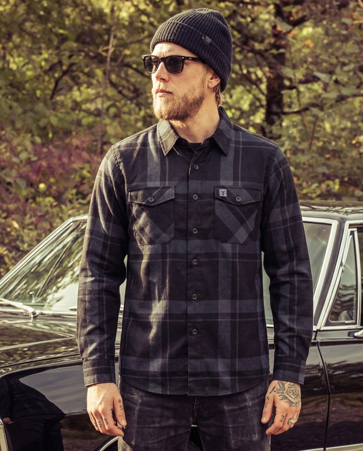 Man wearing a plaid black and gray flannel shirt and sunglasses standing next to a car with trees in the background - The Synister Flannel by Greasy Hands Society