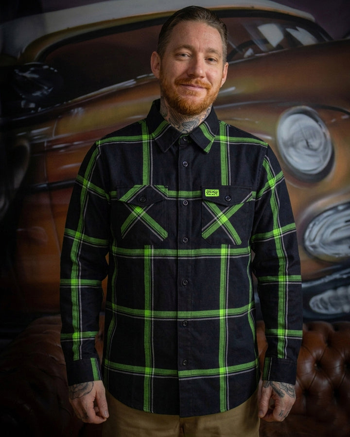 Man wearing a black and green plaid flannel shirt with a vintage car in the background - The Moto Flannel by Greasy Hands Society
