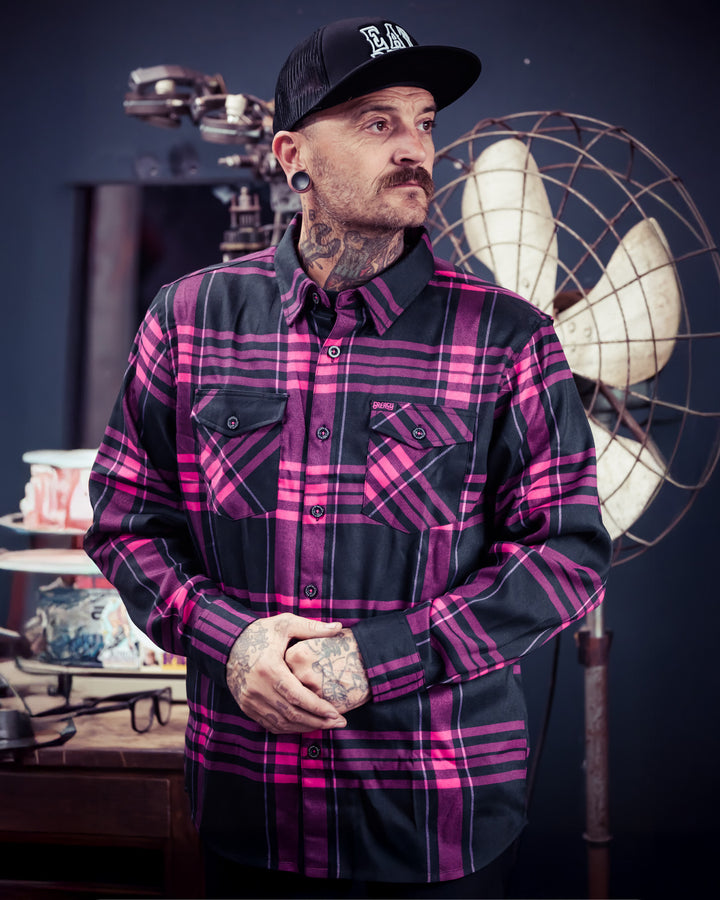 Man wearing a black and pink flannel shirt with a cap, standing in a room with various industrial elements - The Lonely Heart Flannel from Greasy Hands Society