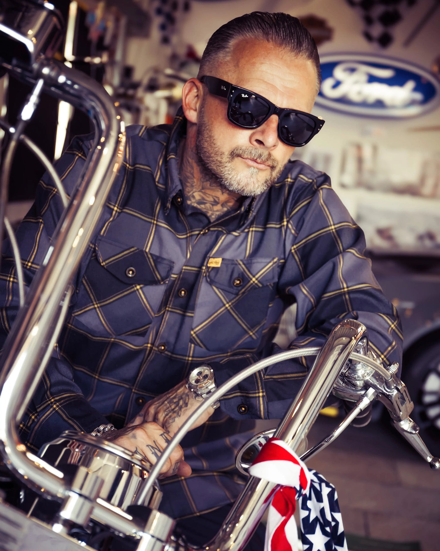 Man wearing sunglasses and a plaid flannel shirt - black, gray and orange flannel shirt - sitting on a motorcycle with a Ford logo in the background - El Lobo flannel by Greasy Hands Society