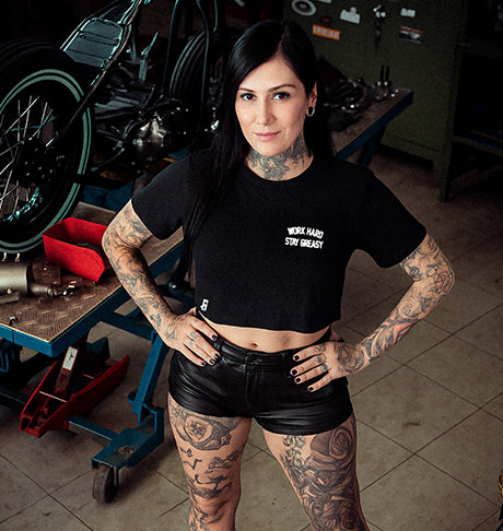 Woman with tattoos wearing a black t-shirt that says work hard stay greasy and shorts in a motorcycle workshop setting