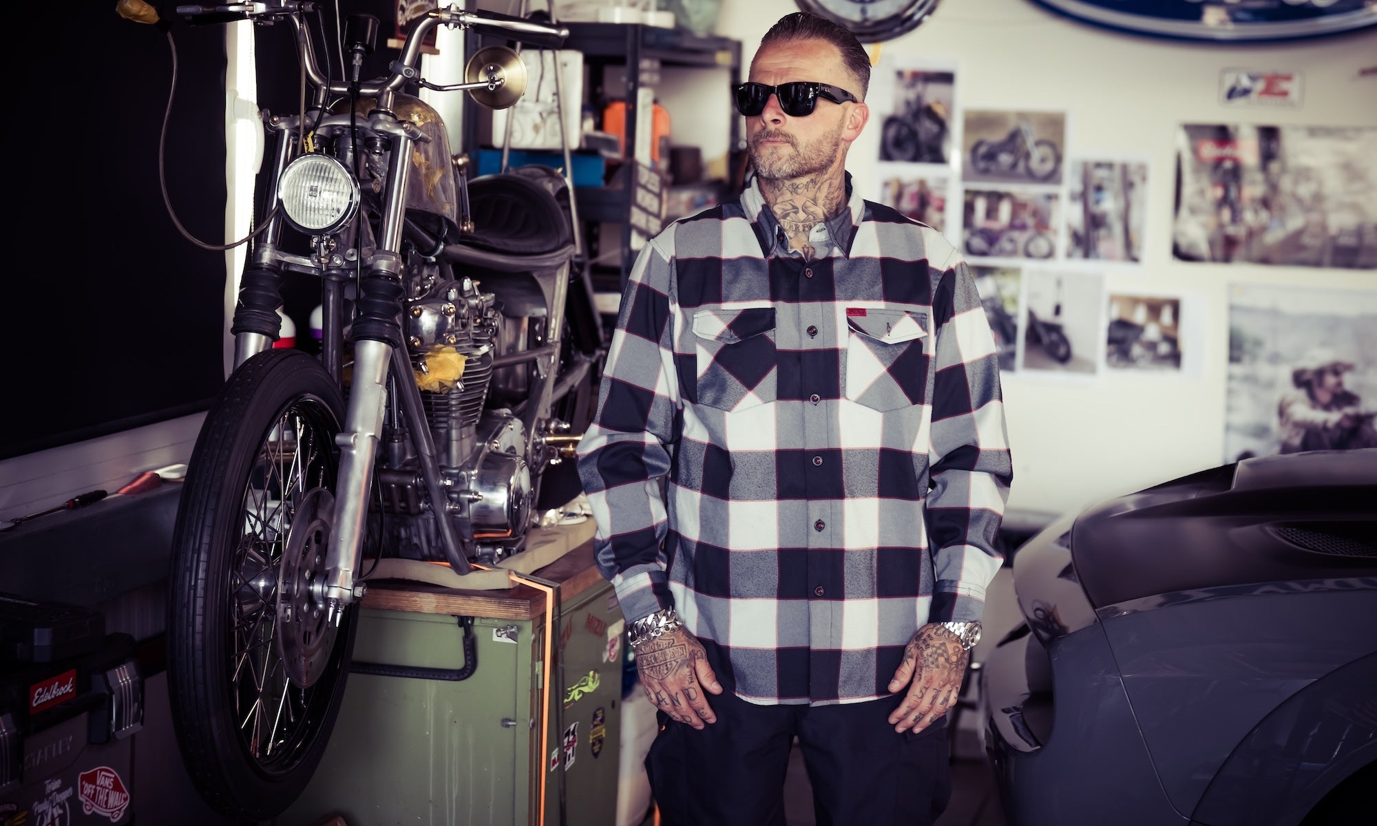 A man wearing a black and white buffalo plaid flannel shirt and sunglasses stands in a garage next to a motorcycle. A Ford logo is visible on the wall behind him.