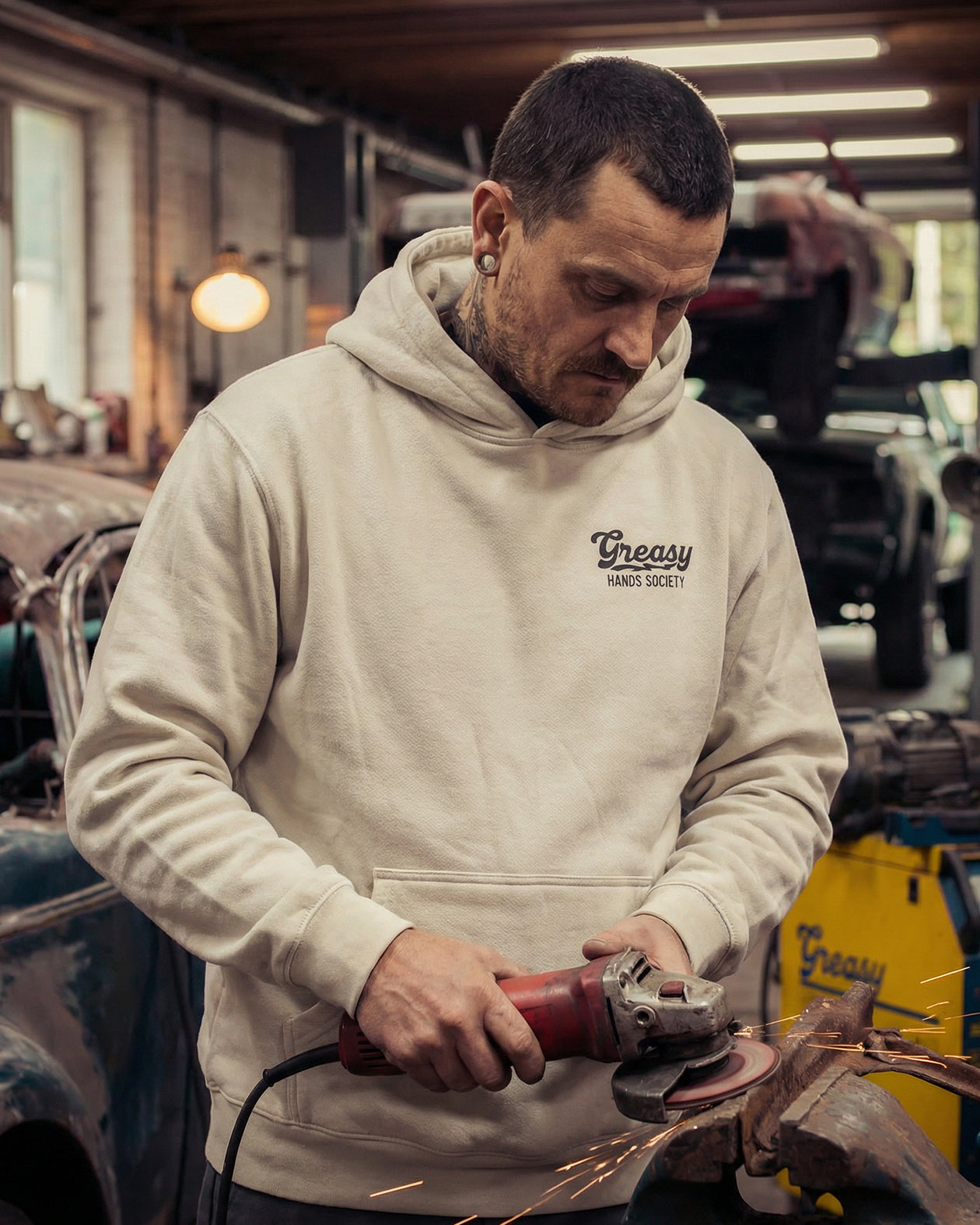 Man wearing a the destination graphic hoodie 'Greasy Hands Society' hoodie using a grinder in a workshop.