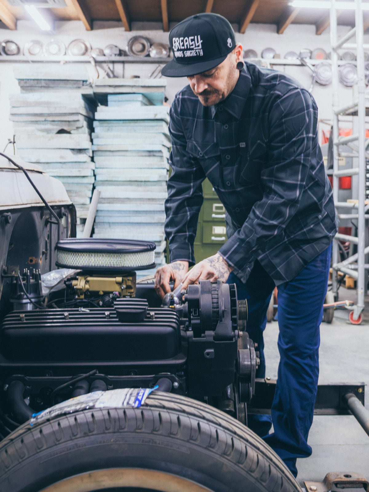 Man working on a vehicle engine in a workshop while wearing a gray plaid shirt - The Dapper Flannel by Greasy Hands Society
