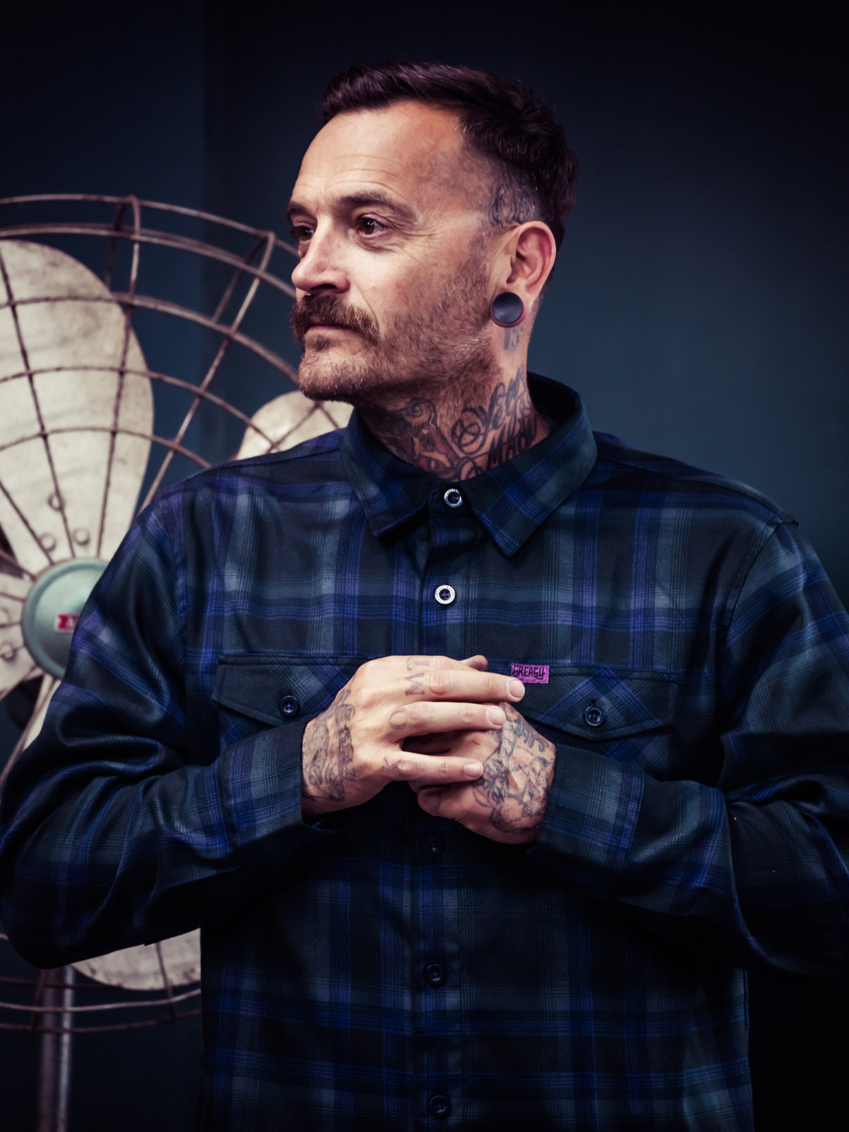 Man wearing a black and purple plaid shirt with tattoos on his arms and neck, standing against a dark background - Kiss the Sky Flannel by Greasy Hands Society