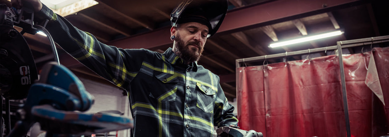 Person in a black and yellow flannel shirt with a welding helmet on with a red curtain in the background - Eat Dust Flannel by Greasy Hands Society