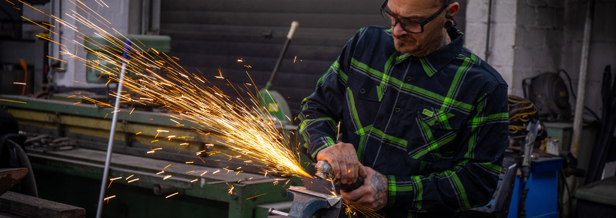 Person wearing a green and black plaid flannel shirt using a grinder with sparks in a workshop setting - The Moto Flannel by Greasy Hands Society