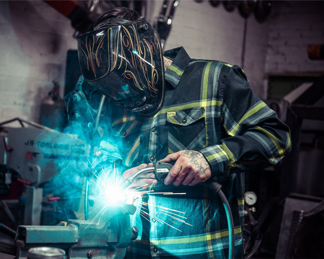 Person welding wearing protective welding helmet and a black and yellow flannel in a workshop setting - Eat Dust Flannel by Greasy Hands Society