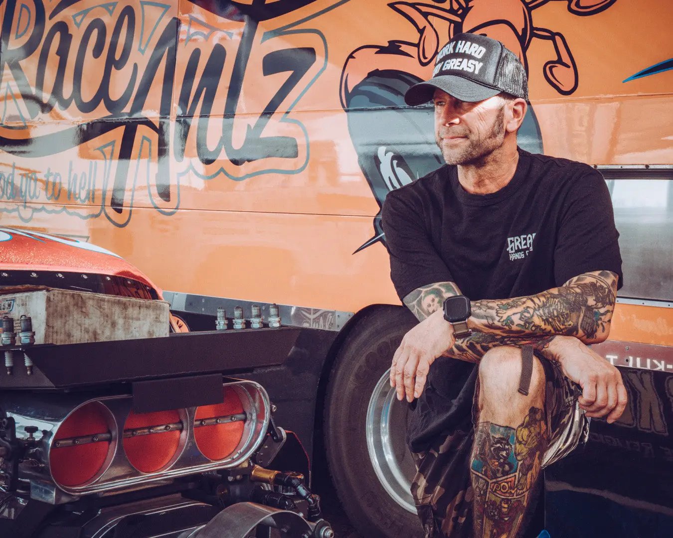 Man sitting on a car with a tattooed arm in front of a branded vehicle - the greasy hands society logo tee by greasy hands society