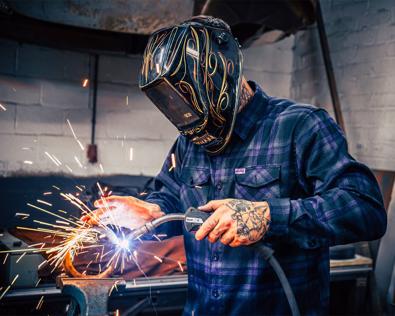 Person welding in a black and purple plaid flannel shirt with sparks flying in a workshop setting - Kiss the Sky Flannel by Greasy Hands Society