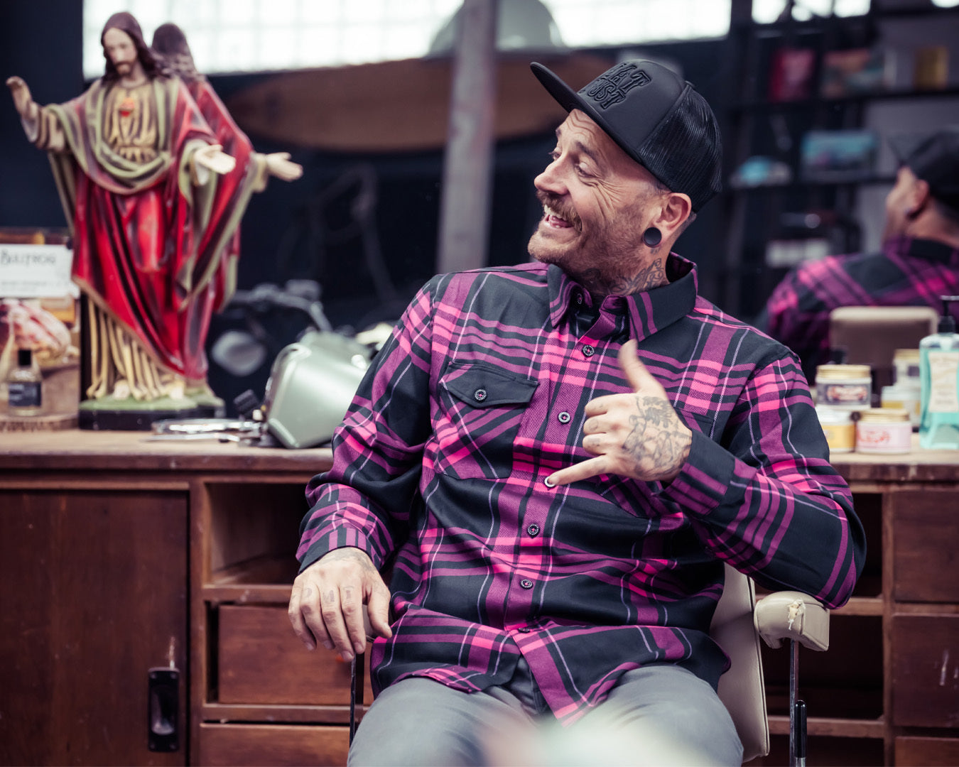 Man in a black and pink plaid shirt sitting in a chair with a statue of a religious figure in the background - Lonely Heart Flannel by Greasy Hands Society