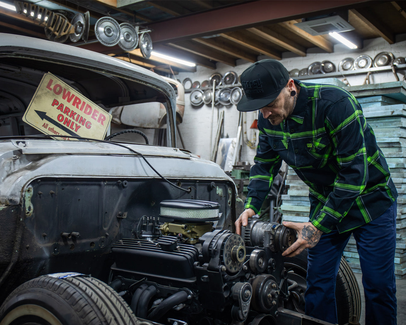 Person in a green and black flannel shirt working on a lowrider car in a garage with a 'Lowrider Parking Only' sign - The Moto Flannel by Greasy Hands Society