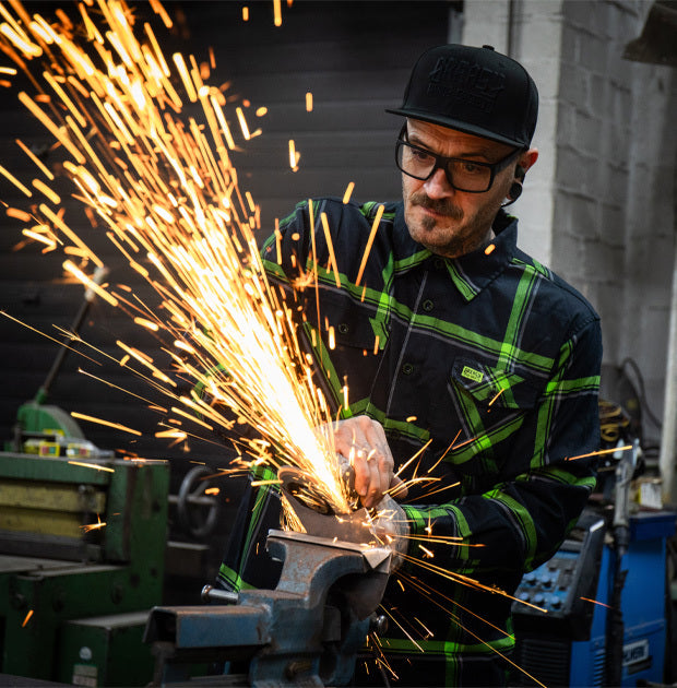 Person wearing a black and green flannel plaid shirt using a grinder with sparks flying in an industrial setting - The Moto Flannel by Greasy Hands Society