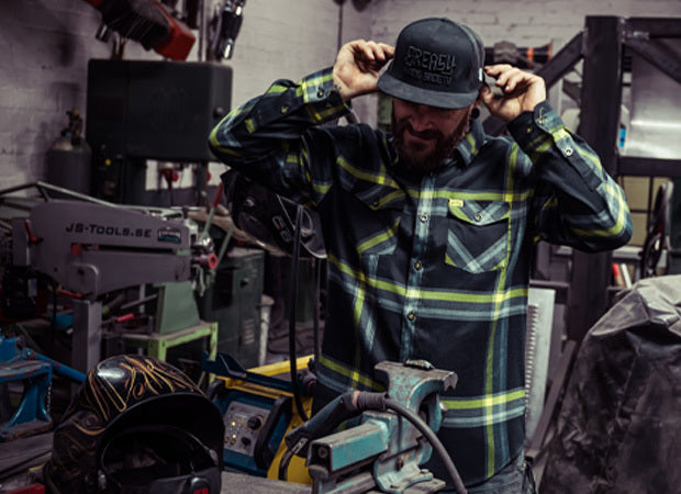 Person wearing a black and yellow flannel shirt and cap in a workshop setting - Eat Dust Flannel by Greasy Hands Society