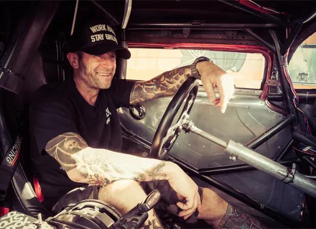 Man sitting inside a vehicle with tattoos on his arms, wearing a cap and black graphic tee shirt - the greasy hands society logo tee by greasy hands society