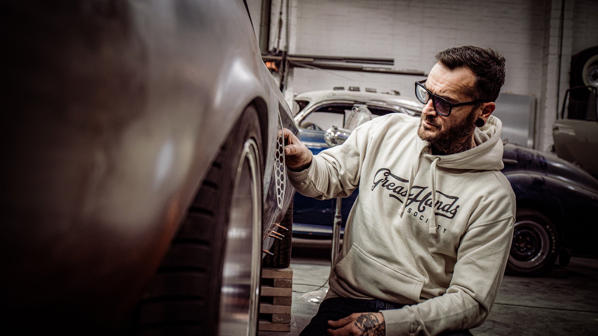 greasy hands society tan script hoodie worn by a man working on his vintage car