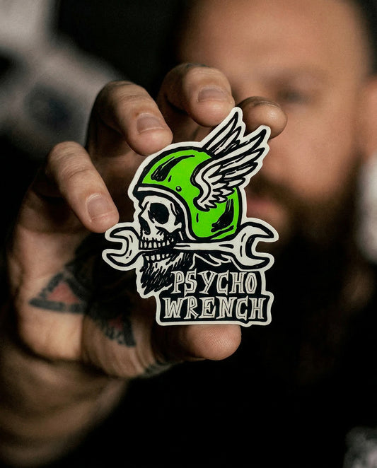 Person holding a 'Psycho Wrench' sticker with a green helmeted skull design.
