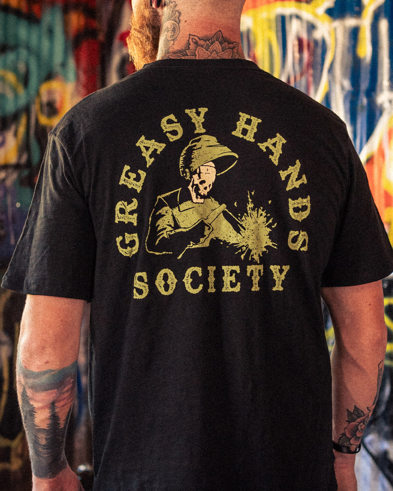 Person wearing a black t-shirt with 'Greasy Hands Society' welding graphic design on the back, standing in front of a colorful mural - Weld On Graphic tee by Greasy Hands Society