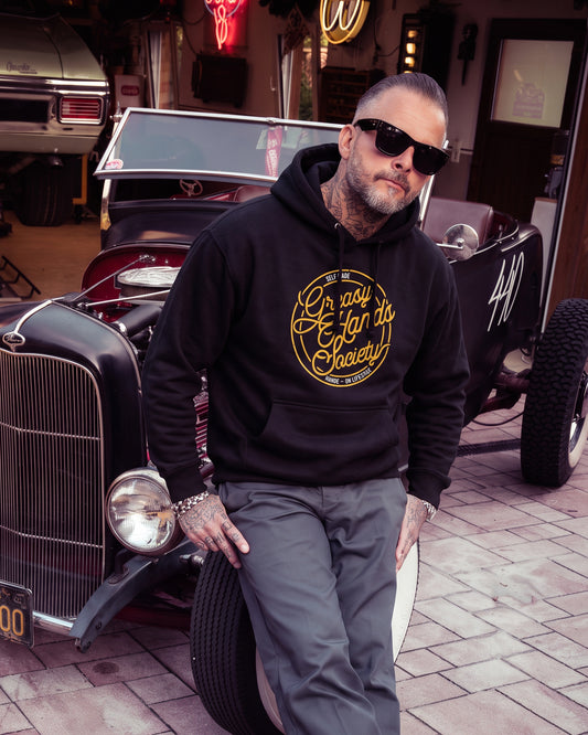 A man wearing a black hoodie with a gold circular graphic that reads 'Greasy Hands Society - Self-Made - Hands-On Lifestyle' and gray pants, leaning against a classic car in a garage setting - Self Made Hoodie by Greasy Hands Society