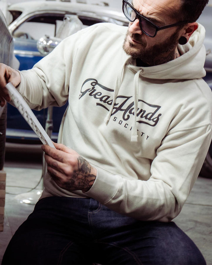 Man wearing a 'Greasy Hands Society' hoodie holding a tool, with a car in the background - Greasy Hands Society Script hoodie in Bone beige