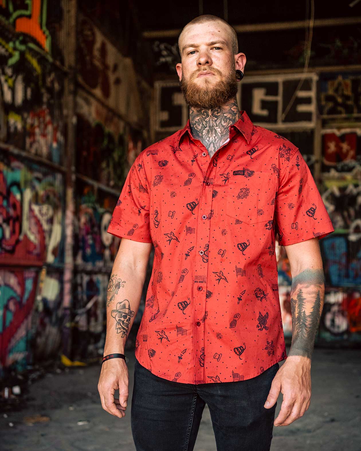 Man wearing a red patterned shirt standing in front of a colorful graffiti wall -an american 250th specia button-up short sleeve shirt - The 1776 button-up short-sleeved shirt by Greasy Hands Society