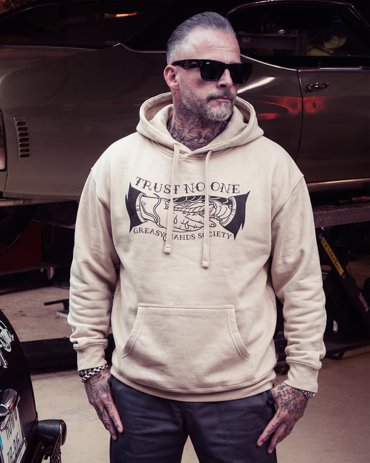 A man wearing a beige hoodie with a black graphic print that reads 'TRUST NO ONE / GREASY HANDS SOCIETY' stands in a garage in front of a car - Trust No One Hoodie by Greasy Hands Society